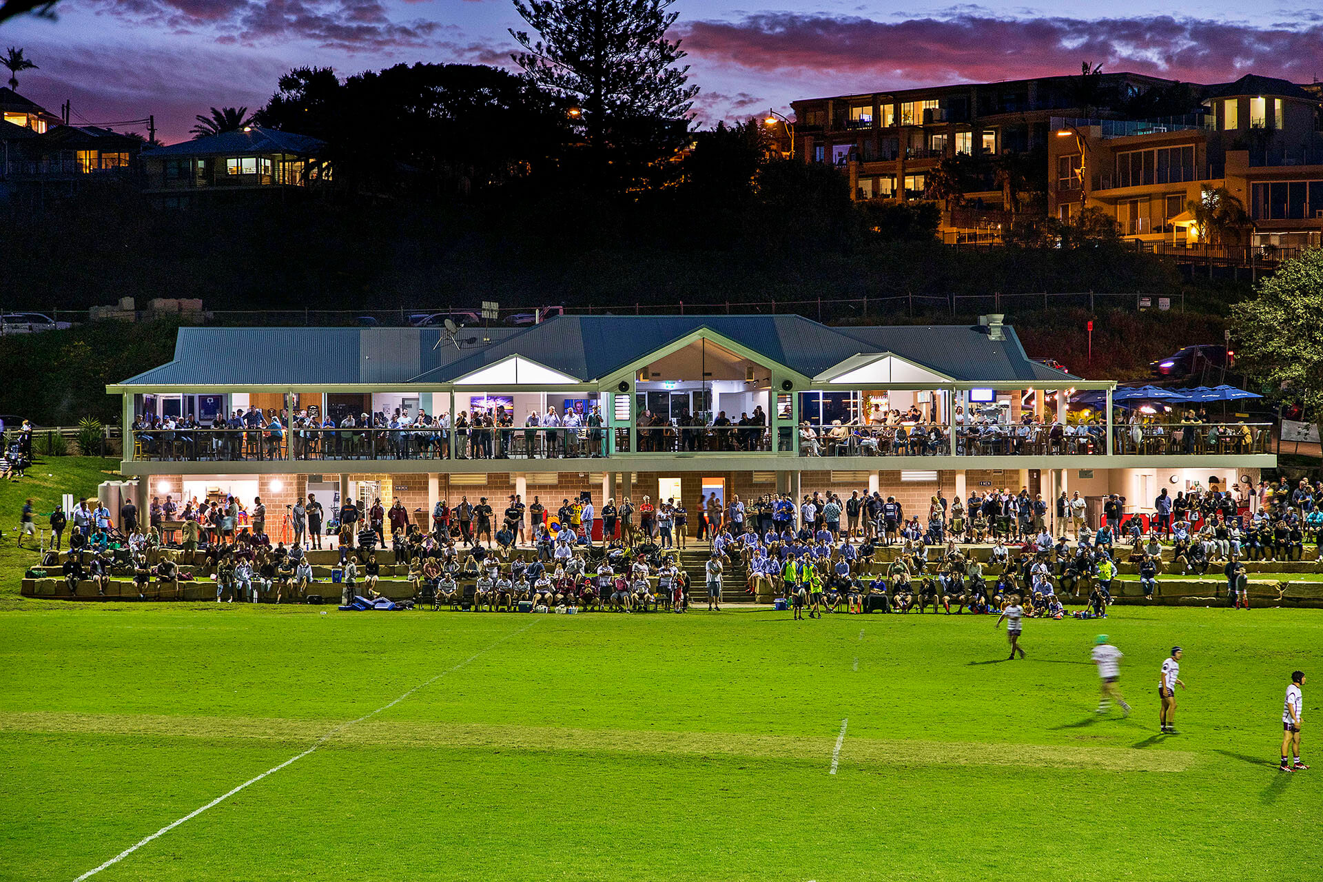 Crowded two-storey building next to large open field with a game of soccer